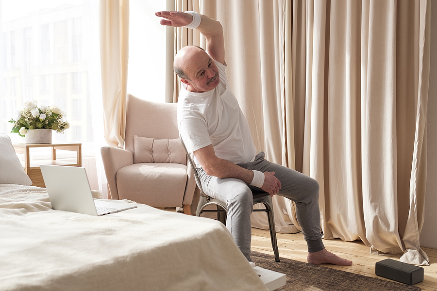 A senior man is seated in a chair and stretching in a bedroom.