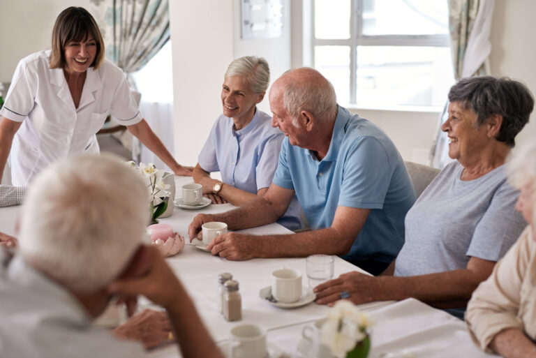 Group of seniors and a nurse laughing and talking together over afternoon tea around a dining table in a retirement home