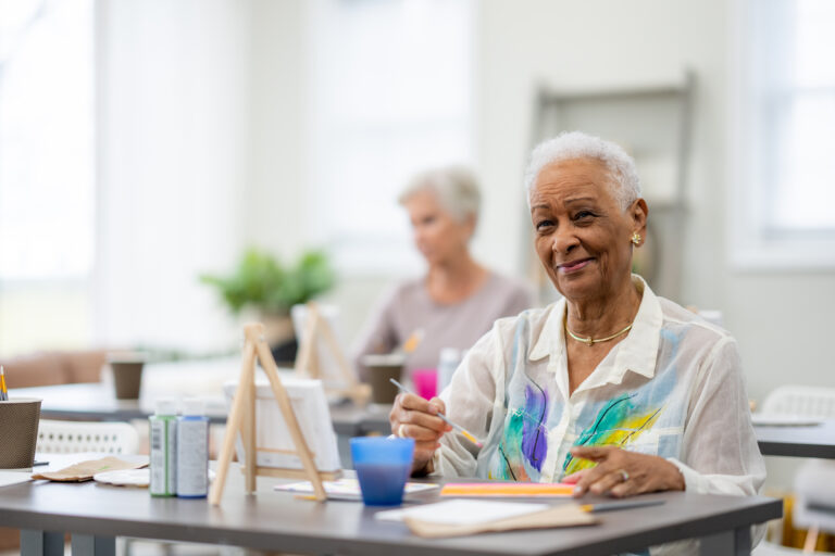 A small group of senior citizens sit together in an art class as they work on individual paintings. The focus is on a woman of African decent in front who is diligently painting away.
