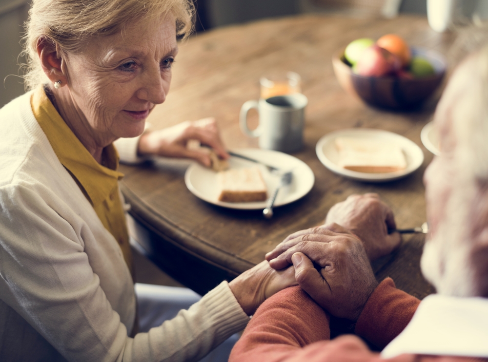 A husband holds the hands of his elderly wife as they eat dinner